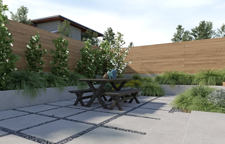 A modern backyard patio featuring a wooden picnic table, concrete pavers with pebbles, and lush green plants
