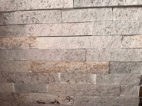 Close-up of stacked stone or tile wall with varying shades of beige and gray, showing texture and grout lines between rectangular tiles